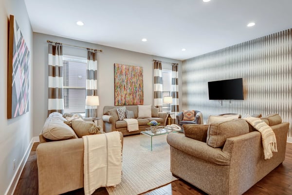 Spacious living room with tan upholstered seating, striped curtains, wall-mounted TV, and colorful abstract artwork