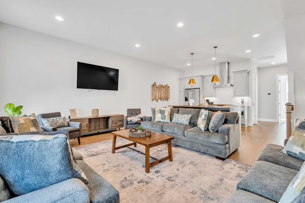 Open-plan living room with gray upholstered seating, wooden coffee table, wall-mounted TV, and kitchen with gold pendant lights visible in background